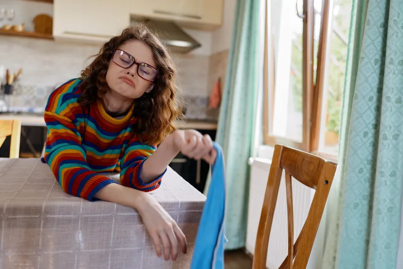 Cheerful woman indoors, casual home scene, colorful striped sweater, glasses, relaxed pose reaching for a bag on the table, warm natural light, cozy atmosphere