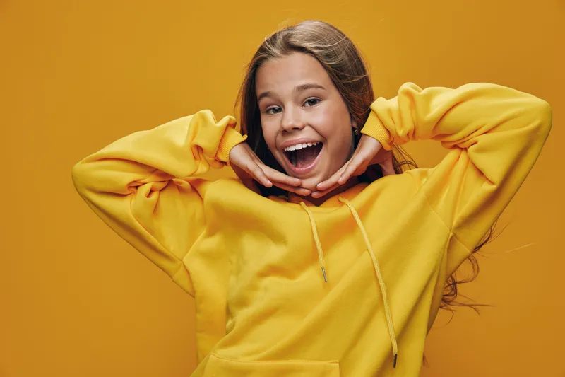 Smiling girl in yellow hoodie posing with hands near face on yellow background. Young female teenager with long hair expressing joy and happiness in casual clothing studio portrait.
