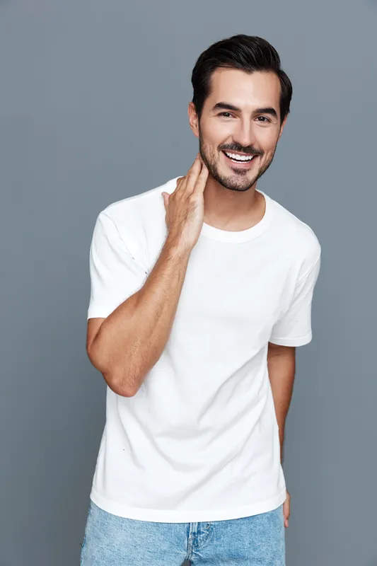 Happy smiling young man with beard wearing a white t-shirt, standing against a solid grey background while touching his face in a relaxed pose.