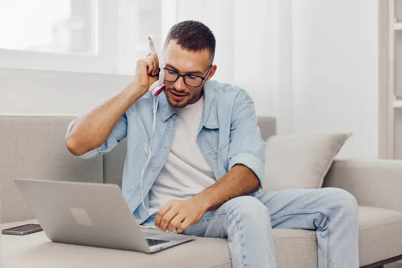Focused man talking on the phone while using a laptop, dressed casually in a cozy modern living room. Engaging in work or study, emphasizing productivity.