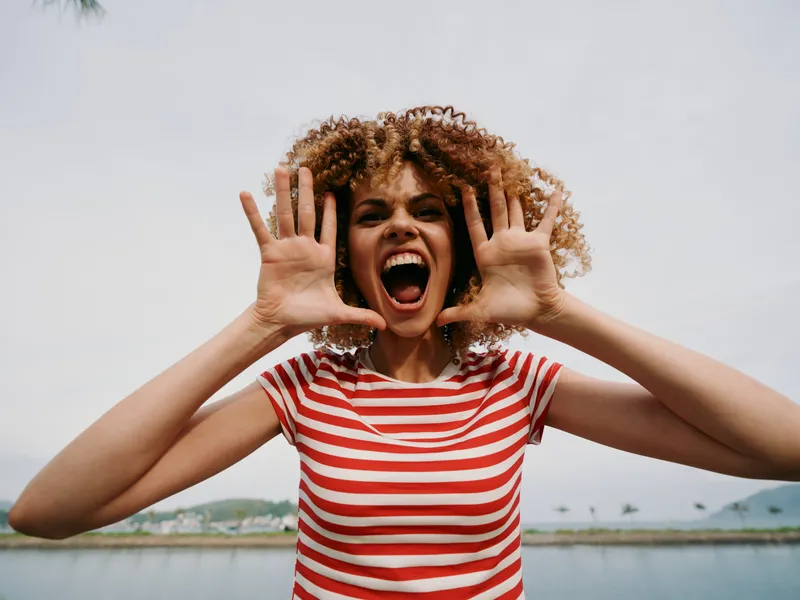 Vibrant portrait of a joyful young woman with curly hair, wearing a red striped tee, cupping hands to her face and shouting with energy against a bright outdoor backdrop.