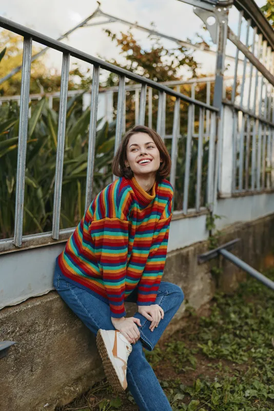 Woman in her late twenties wearing a colorful striped sweater and jeans, smiling while sitting on a garden wall by a metal fence, casual outfit with relaxed lifestyle travel vibe.