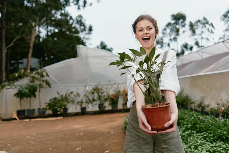 Young woman joyfully holding a potted plant in a greenhouse, surrounded by greenery and soft natural light, embodying a passion for gardening and nature
