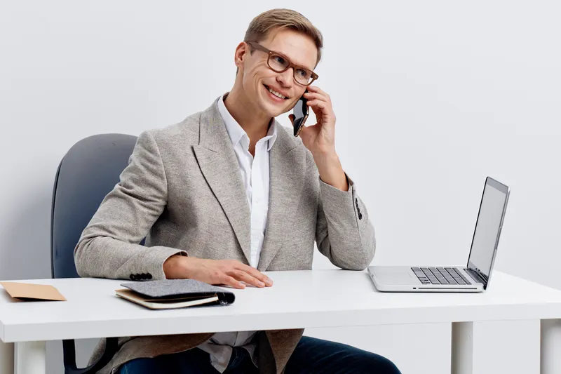 Professional man with glasses in business attire smiling and talking on the phone while sitting at a desk with a laptop, notebooks, and documents, in a modern office environment, concept of communicat