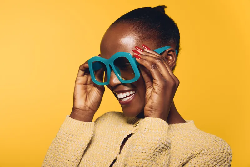 Smiling African woman with dark skin adjusting large blue sunglasses against yellow background. Concept of fashion, summer style, beauty, and joyful mood in studio portrait.