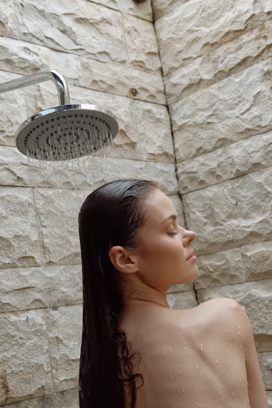 woman showering outdoors with wet hair, natural stone wall background, relaxed expression, wellness and self-care moment in natural light