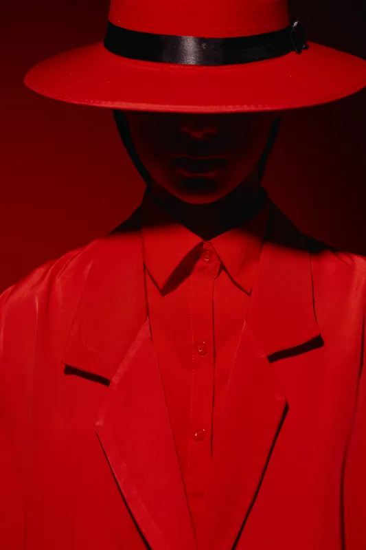 A mysterious person in a red wide-brimmed hat and red blazer is captured under dramatic lighting. Shadows obscure the face creating a bold beauty and fashion concept with creative style.