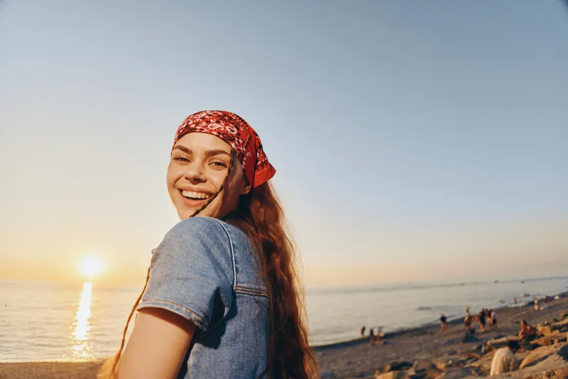 smiling woman wearing a red bandana and denim jacket enjoys a sunny beach sunset with warm light and relaxed atmosphere outdoors by the sea