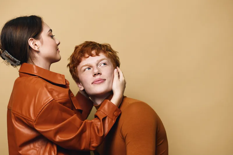 Young couple with red hair and brown jackets posing against beige background. Woman gently holds man face with affection and care in close intimate moment.
