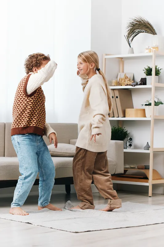 Two children dance indoors, barefoot and laughing, as they twirl in cozy sweaters and jeans, creating a joyful moment of playful movement and connection.