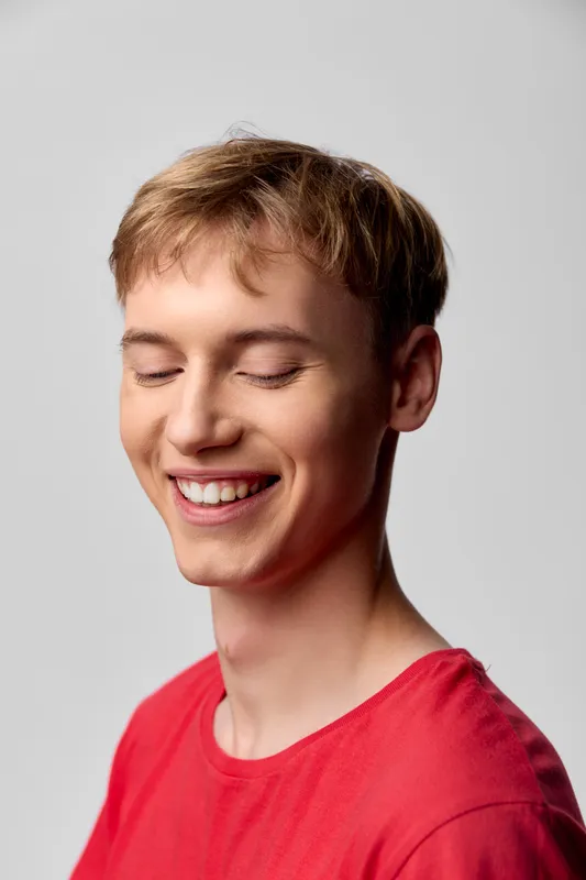 A smiling man with closed eyes wears a red shirt in a bright studio, conveying joy, calm and relaxed expression through soft lighting and candid mood
