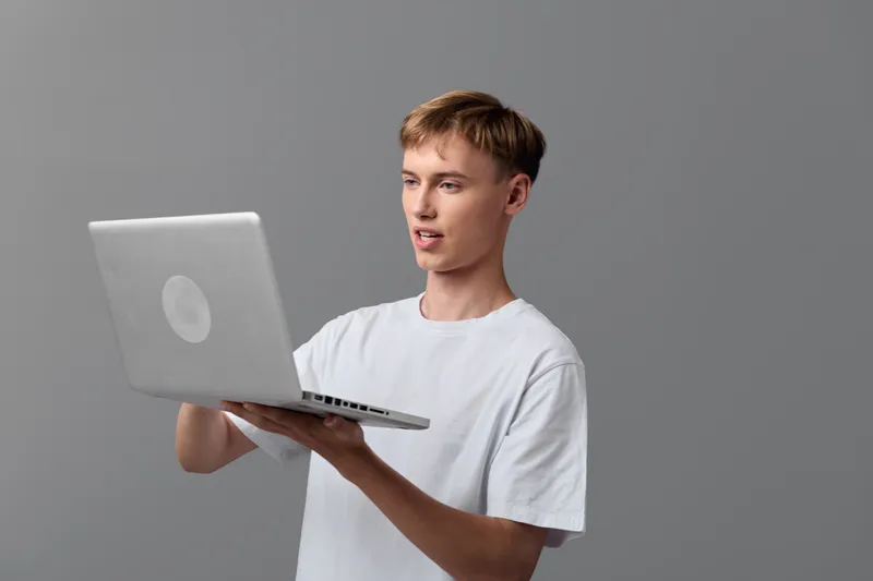 Laptop, man holding device for technology use in a casual studio setting, remote work and education visuals with focused expression and clean gray background, modern minimal setup