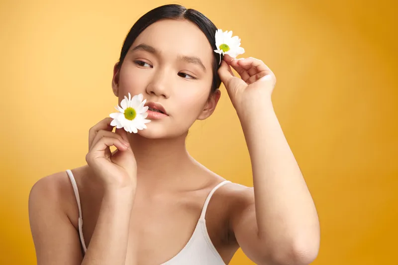 Asian woman with delicate features holds white flowers near her face against a warm yellow backdrop, creating a soft, fresh portrait with a calm, inviting atmosphere.