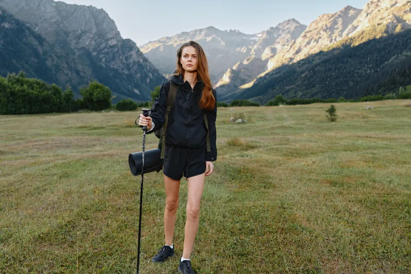 A young woman wearing hiking gear stands in a grassy valley with a trekking pole, surrounded by wide open meadow and dramatic mountains under a clear sky, conveying adventure and exploration.