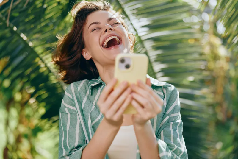 Woman, phone, outdoors, laughter, joyful, smiling, happy, portrait, nature, greenery, background, sunlight, sunny, candid, moment, joy, connection, media, app, social, life, leisure, fashion, casual,