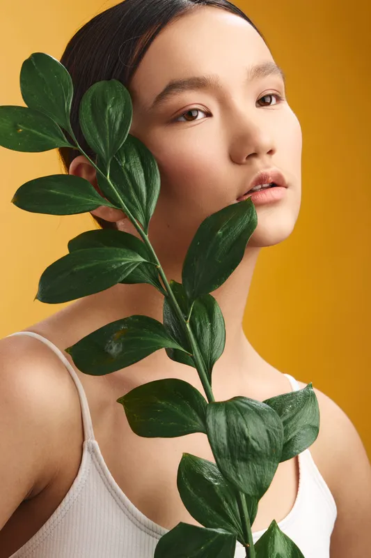 asian woman holds a leafy stem against a warm yellow backdrop, presenting a natural portrait that conveys calm confidence, modern style, and subtle elegance in a studio setting.