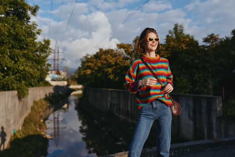 Woman in a colorful striped sweater and sunglasses, smiling and holding a coffee by an urban canal in jeans; casual outfit, autumn light, travel lifestyle and relaxed confident pose.