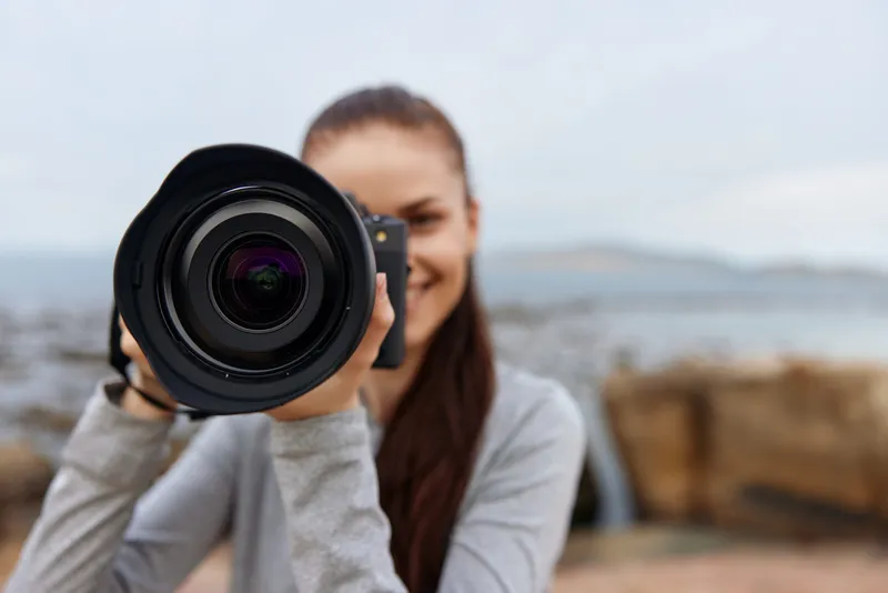 Young woman photographer smiling while holding a professional camera against a scenic outdoor background, showcasing enthusiasm for photography