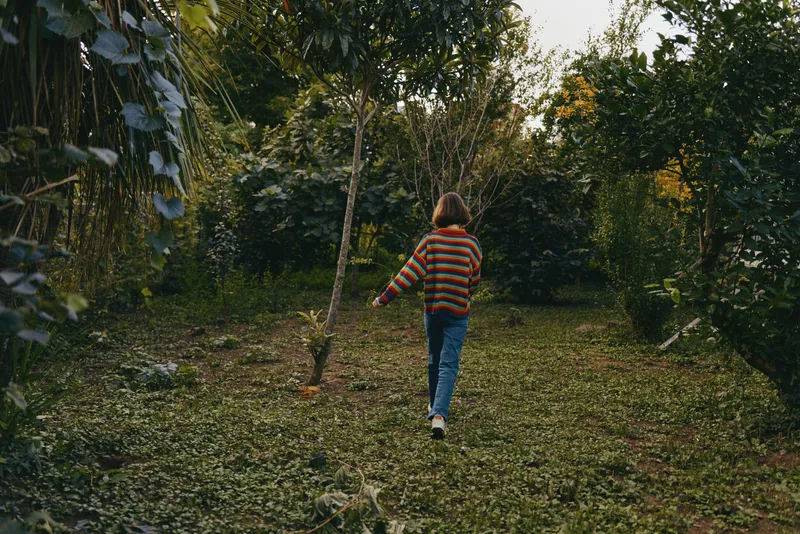 Young woman striped sweater walking in a tropical outdoor garden, casual outfit with jeans and sneakers, exploring a forest path, relaxed lifestyle travel scene amid lush nature.