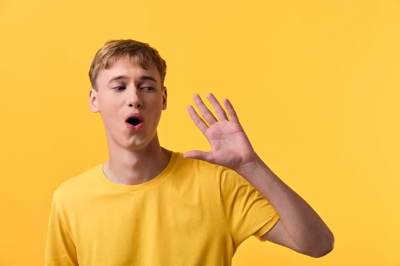 Surprised expression and raised hand against a bright yellow background, signaling stop, alertness, and vocal emotion in a studio shot. Youthful energy and dynamic gesture convey urgency and attention