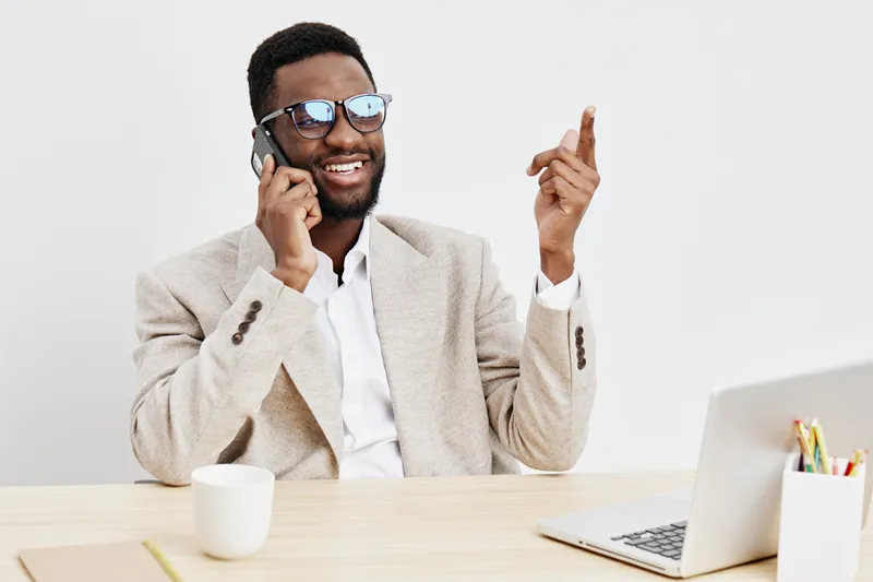 Smiling man with dark skin, wearing glasses and a beige blazer, talking on mobile phone while sitting at a desk with a white mug, laptop, and colorful stationery, isolated on plain white background, b