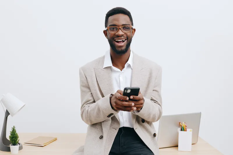 Confident happy businessman standing at desk using smartphone, smiling, casual blazer, white shirt, modern workspace, clean background, lifestyle concept
