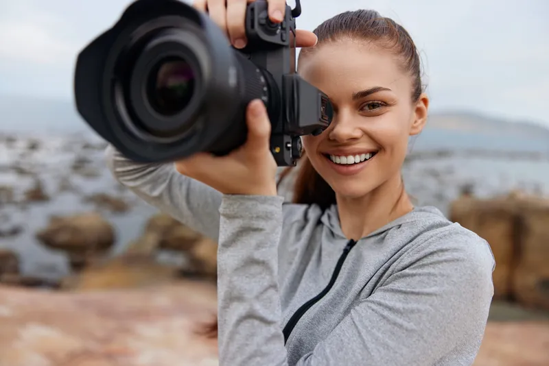 Happy young woman with long hair in a gray hoodie holding a camera, smiling outdoors by the seaside, showcasing her enthusiasm for photography and nature