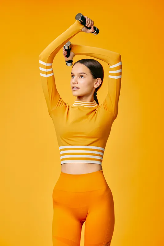 Woman fitness portrait: fit young woman in orange activewear lifting small dumbbells overhead in a sport training pose, highlighting toned core, focused expression and vibrant studio background.
