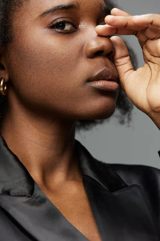 Elegant young Black woman in a sleek black jacket, expressing introspection with a thoughtful pose against a muted gray background