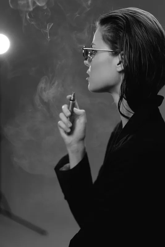 Black and white profile of a woman smoking, wearing glasses and dark clothing in a moody atmosphere.