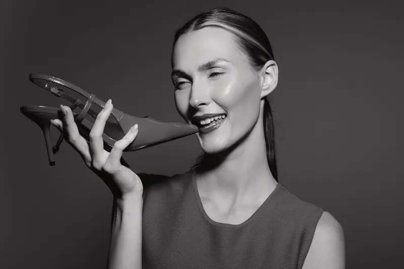 Slim white woman with slicked hair wearing sleeveless top holds high heel shoe near smiling face in studio. Concept of fashion, style, and elegance.