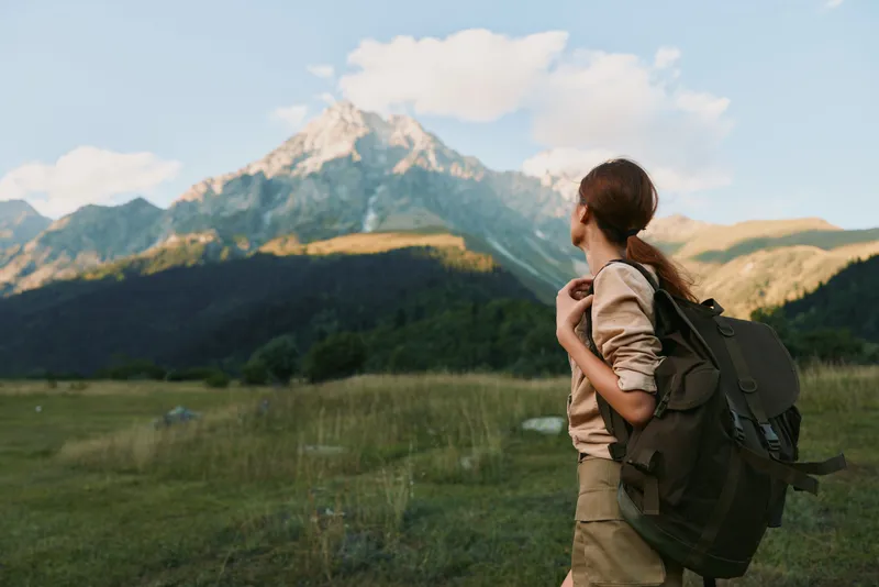 Woman hiking outdoors with a backpack, looking toward a distant mountain peak in a sunlit valley, capturing a moment of nature, adventure, and calm scenery