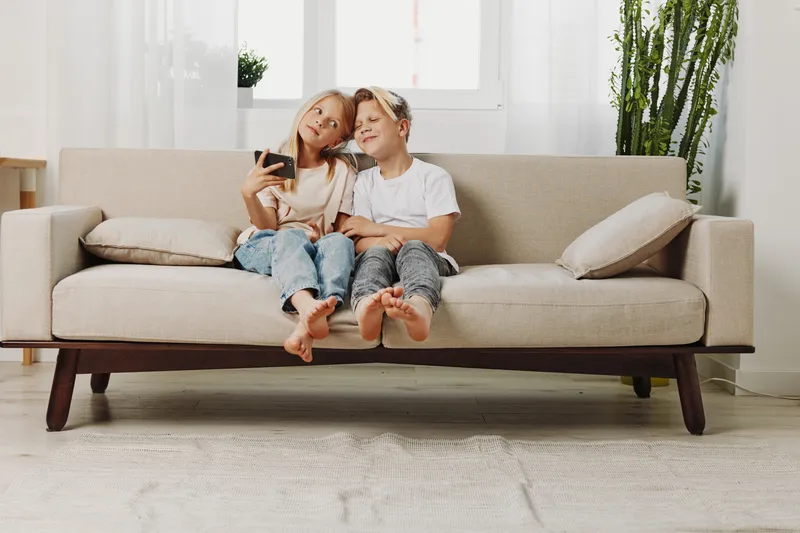 Two happy children sit barefoot on a modern beige sofa in a bright living room. They smile and take a selfie with a smartphone, enjoying family time and childhood moments.