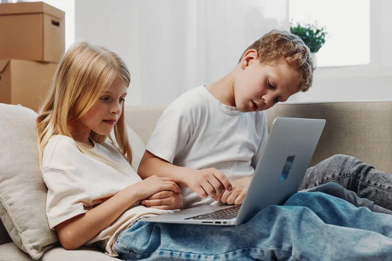 Two children in casual clothes use a laptop on a sofa in a bright room with natural light. Concept of learning, technology, and childhood curiosity.