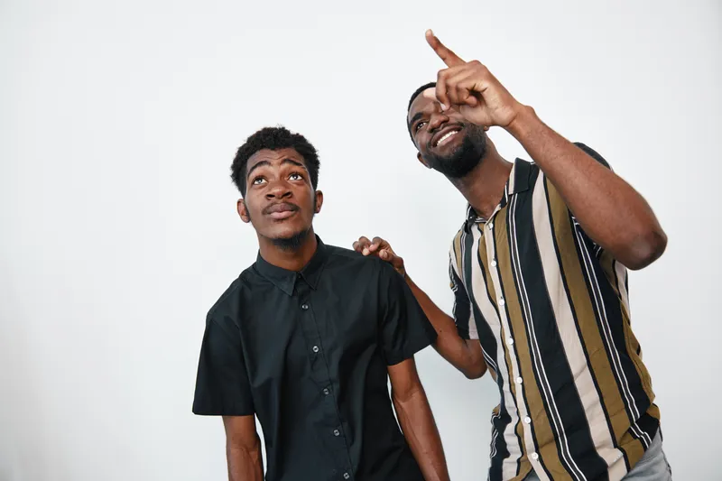 Two African American men with dark skin standing together on white background, one pointing up and the other looking in the same direction with casual clothing and friendly expressions.