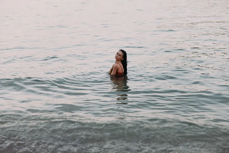A lone woman stands waist deep in calm sea water, facing the shore with a serene gaze, gentle light on rippling waves creating a tranquil beach mood and quiet horizon.