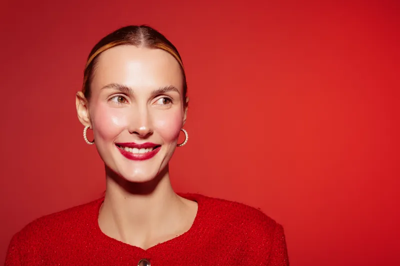 Smiling woman with red lipstick and earrings looks away on red background. Portrait of happy female model with natural skin and bright makeup. Concept of beauty and fashion.