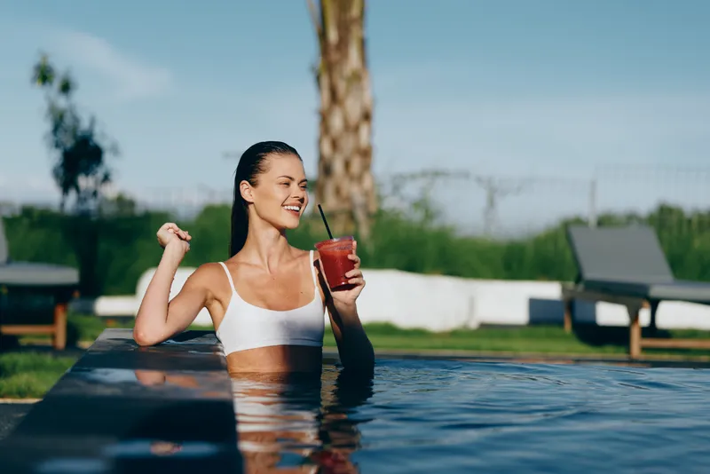Smiling woman in a white swimsuit relaxes in an outdoor pool, sipping a smoothie beside palm trees. Sunlit water and garden scenery create a wellness, relaxation, lifestyle mood.