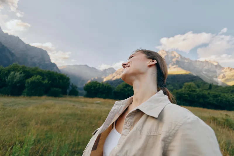 A woman in a white shirt stands in a wide field, mountains in the distance, looking up at the sky. A lifestyle moment conveying nature, calm, and outdoor freedom.