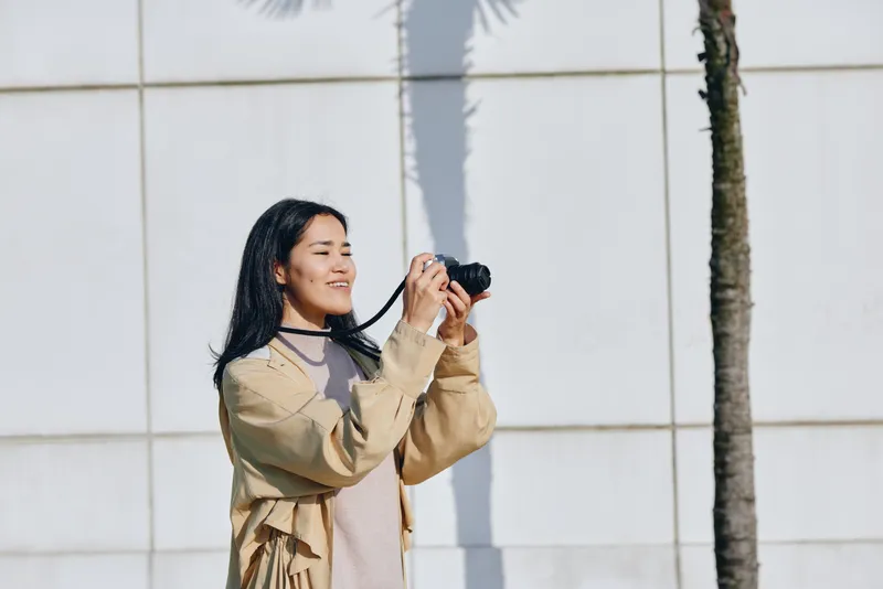 Woman holding camera outdoors, smiling beside a palm tree and modern wall, casual fashion vibe, candid street style photography for lifestyle concepts on a sunny day.