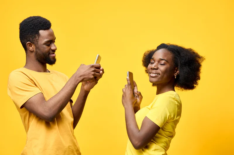 Two young African models using smartphones against a bright yellow background, expressing joy and connection with each other