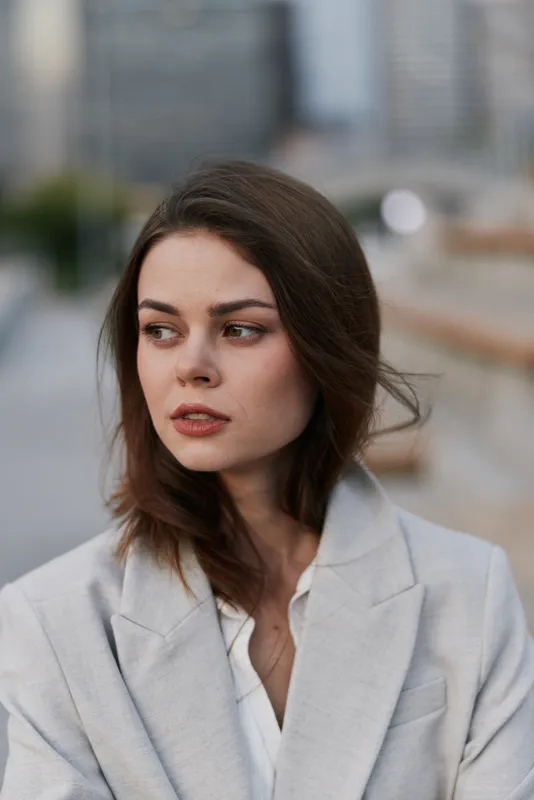 Woman in light gray jacket with thoughtful expression looking aside in urban outdoor setting, natural light, soft color palette, autumn fashion portrait