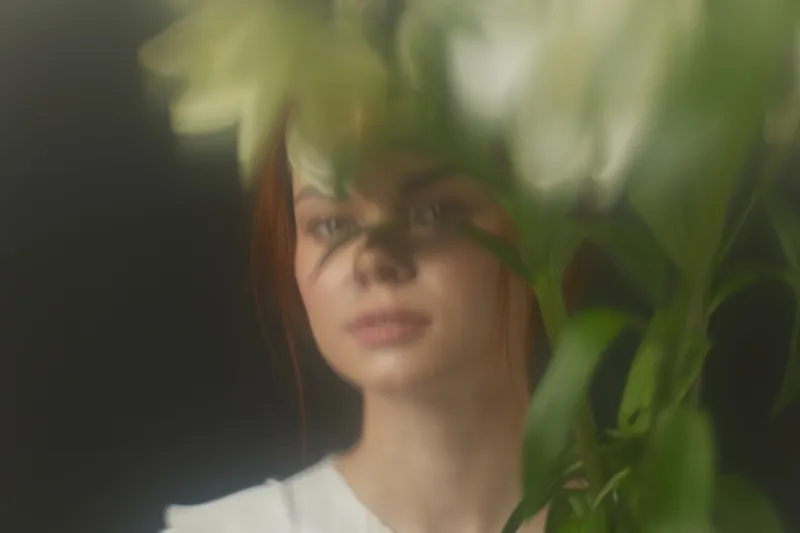 Ethereal portrait of a young woman with reddish hair, partially obscured by flowers, embodying serenity and natural beauty in a softly lit environment