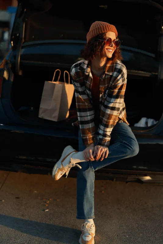 Woman shopping by a car trunk at sunset, smiling in sunglasses and beanie. Casual denim and sneakers, candid lifestyle portrait with authenticity, golden hour glow, mindful living and emotional