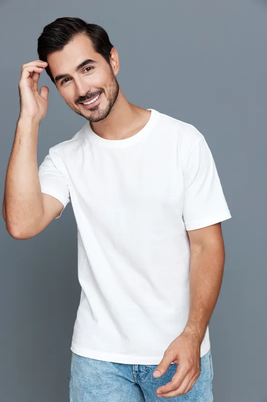 Happy smiling man wearing a white t-shirt with a friendly pose against a plain grey background, showcasing confidence and casual lifestyle.