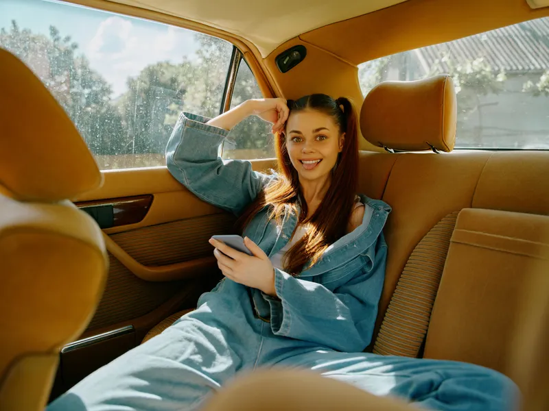 Happy young woman sitting in a car, wearing a denim jumpsuit, smiling while using a smartphone, conveying joy and casual style.