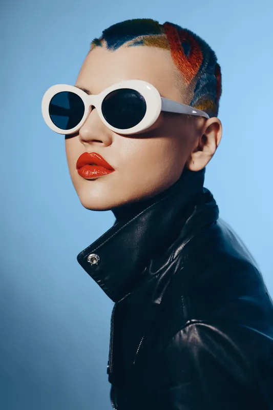 Young woman with colorful braided hair wearing white sunglasses and glossy red lipstick in a black leather jacket, modern fashion style, studio lighting.