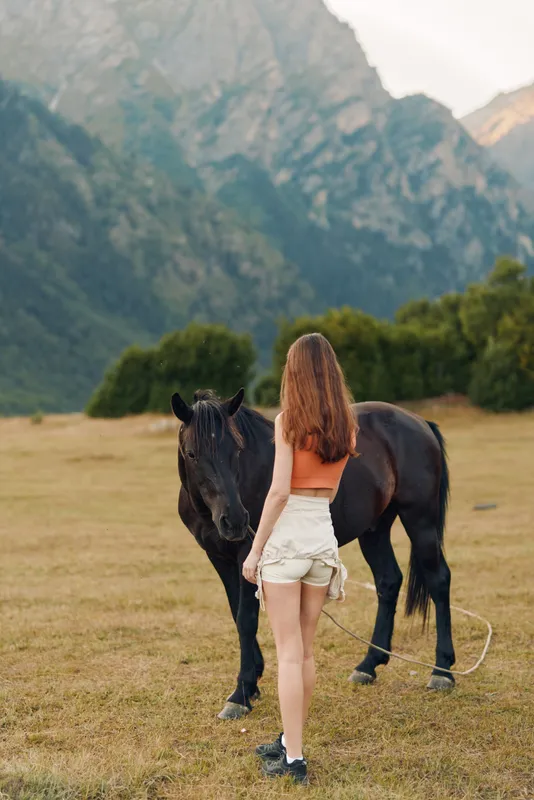 A young woman stands in a wide meadow with a dark horse beside her, mountains rising behind them. The scene conveys calm companionship, outdoor freedom, and rustic charm.