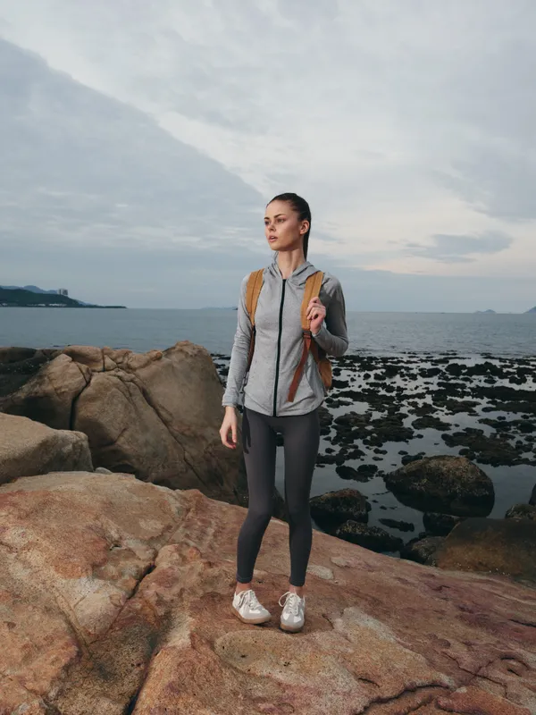 Young female model in athletic wear standing on rocky shore, embodying outdoor adventure and fitness with a serene ocean background