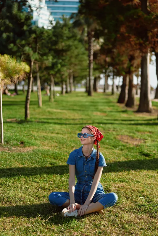 Sitting woman in denim jumpsuit and red headscarf relaxing outdoors on green grass under bright sunny sky in urban park with trees background